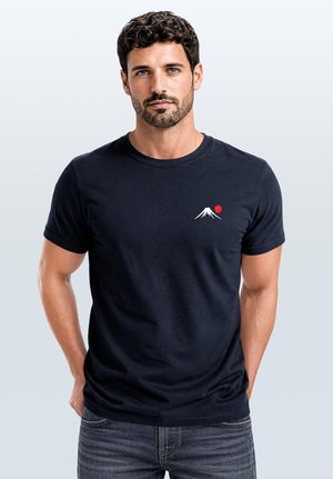 Man with short curly hair and beard wearing a black t-shirt with a small white mountain and red sun graphic on the chest.