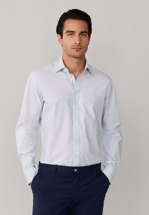 Man die een lichtblauw overhemd met patroon en lange mouwen draagt, gecombineerd met marineblauwe broek, staand met handen in de zakken tegen een lichtgrijze achtergrond.