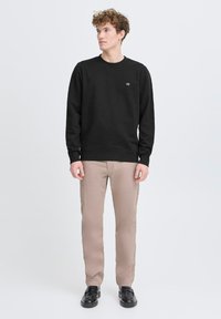 Felpa nera con scollatura a girocollo, polsini e orlo a coste, che presenta un piccolo logo. Abbinata a pantaloni beige chiaro e scarpe nere.