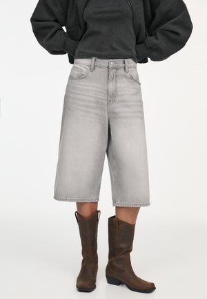 Personne portant un short en jean gris clair longueur genou, un haut foncé et des bottes de cowboy marron, debout devant un fond uni.