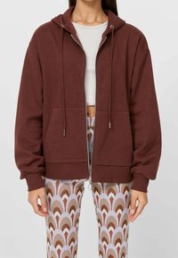 Bruine zip-up hoodie van zacht materiaal, met een capuchon met trekkoord, voorzakken en elastische manchetten. Gecombineerd met patroonleggings.