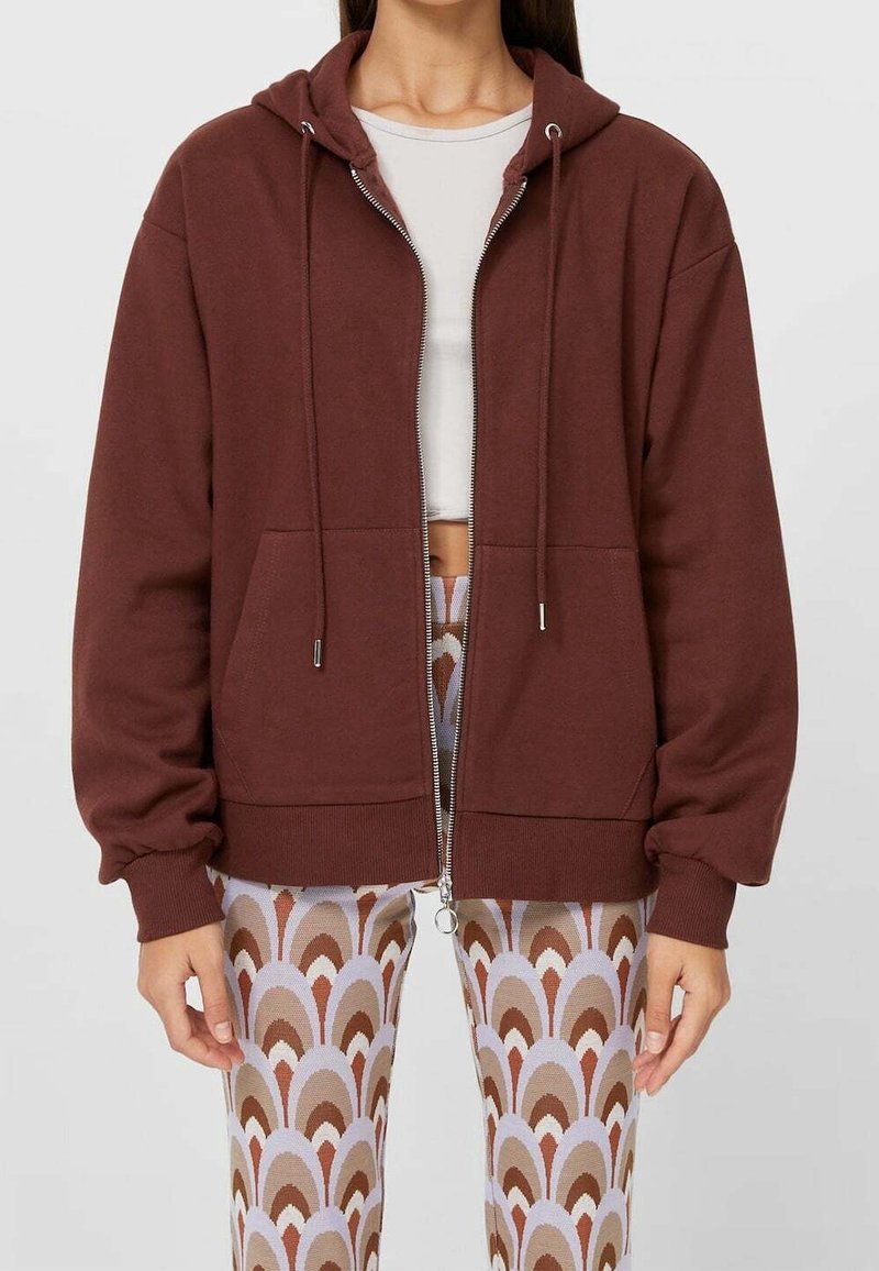 Bruine zip-up hoodie van zacht materiaal, met een capuchon met trekkoord, voorzakken en elastische manchetten. Gecombineerd met patroonleggings.