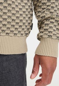 Suéter de punto beige con un patrón de cuadros texturizado, con acentos en verde oscuro. Puños y dobladillo acanalados. Etiqueta con la inscripción "Blend" visible.