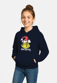 Una giovane donna sorridente con i capelli raccolti in un chignon indossa una felpa blu navy con il volto del Grinch e un cappello di Babbo Natale, accompagnata dalla scritta "Sentendosi Grinch."