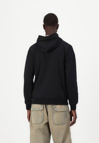 Čierna mikina s kapucňou a voľným strihom, obsahujúca klokanie vrecko a rebrované manžety. Nosená s svetlými cargo nohavicami.