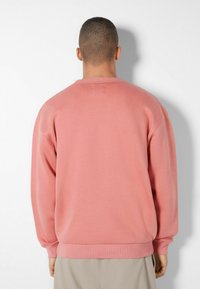 Man med kort hår som bär en enkel rosa sweatshirts och beige byxor, vänd mot en vit vägg med en skugga kastad bakom honom.