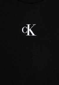 T-shirt de algodão preta com um logótipo "cK" branco no canto superior esquerdo. Decote redondo e textura suave.