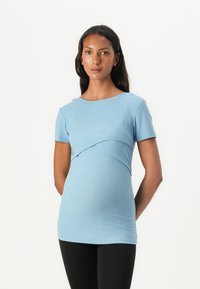 Lyseblå ribbet skjorte med korte ermer, med et overlapping design foran. Brukt over svarte leggings, står modellen mot en ensfarget bakgrunn.