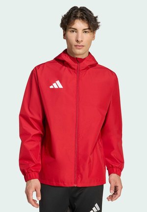 Giovane uomo che indossa una giacca sportiva rossa con zip e cappuccio, con un logo bianco sul petto, abbinata a pantaloni neri con un logo bianco.