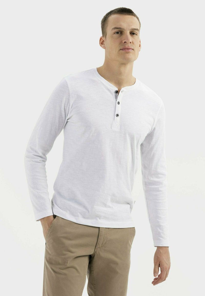 Weißes Langarmhemd mit Henley-Ausschnitt, drei schwarzen Knöpfen und weicher Textur, kombiniert mit khakifarbenen Hosen.