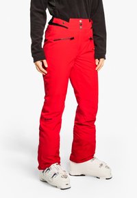 Rote Skihose mit glatter Textur, mit Knopf- und Reißverschluss, seitlichen Taschen und einem Logo am linken Bein. Getragen mit weißen Skischuhen.