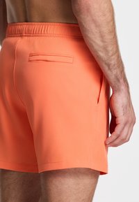 Oranžinės maudymosi šortai su elastinga juosta, šoninėmis kišenėmis ir užpakaline kišene, pagaminti iš lygaus, lengvo audinio.