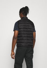 Schwarze, gepolsterte Weste mit hohem Kragen, die vertikale Nähte und eine glatte Textur aufweist, getragen über einem marineblauen Kurzarmshirt und dunkler Hose.