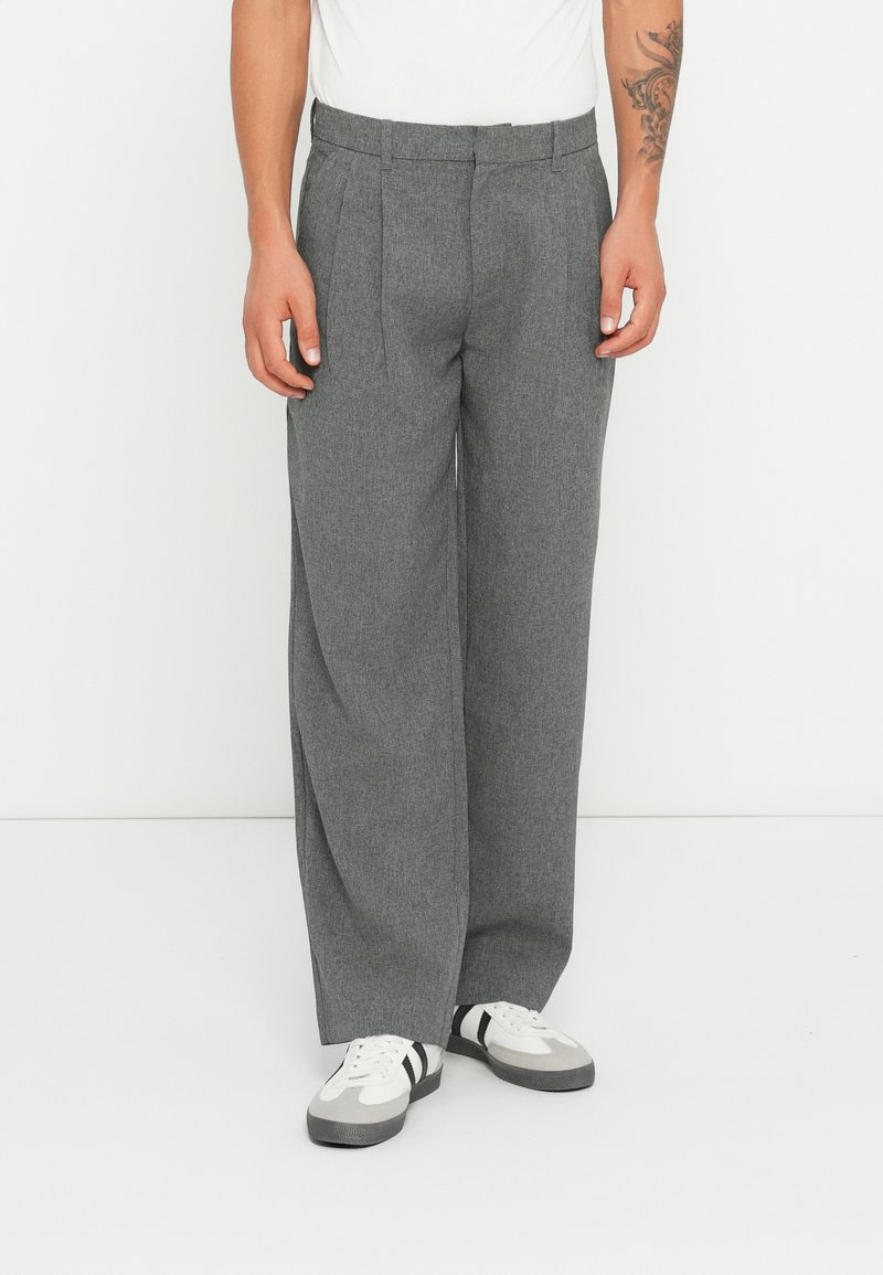Graue, weit geschnittene Hose mit strukturiertem Muster, Falten vorne und Gürtelschlaufen, kombiniert mit weißen Sneakern mit schwarzen Streifen.
