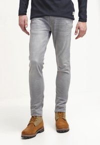 Harmaat slim-fit farkut, joissa on sileä pinta, yhdistettynä ruskeisiin ja vihreisiin nilkkasaappaisiin, joissa on keltaiset nauhat ja karkeat pohjat.