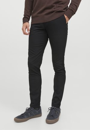 Mand iført sort slim-fit bukser med hænderne i lommerne og mørkegrå sneakers med brune såler.