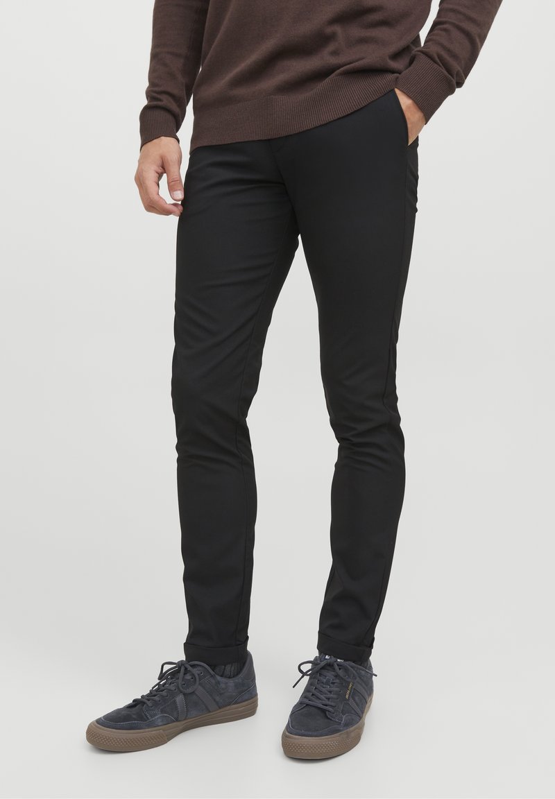 Homme portant un pantalon noir slim, les mains dans les poches et des baskets gris foncé avec des semelles marron.