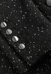 Czarna teksturowana tkanina z białymi drobinkami ma w przekątnej rzędzie pięć dużych, błyszczących, okrągłych srebrnych guzików.