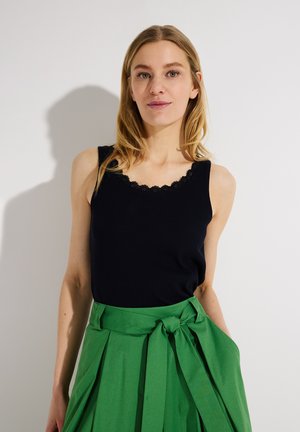 Frau mit blondem Haar, die ein ärmelloses Oberteil mit schwarzer Spitzenborte und einen hoch taillierten grünen Rock mit gebundenem Gürtel trägt, steht vor einem schlichten Hintergrund.