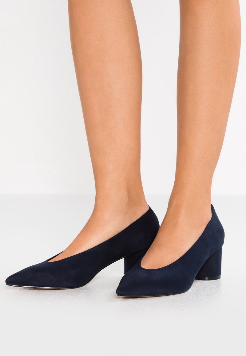 Femme portant des chaussures en daim bleu marine à talon bas bloc avec des bouts pointus sur une surface blanche.
