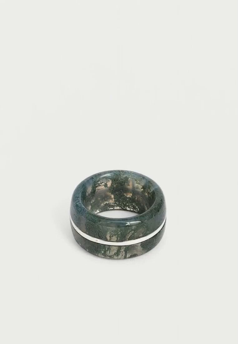 Une large bague vert foncé à l’aspect marbré, ornée d’une fine bande argentée, repose sur une surface claire.