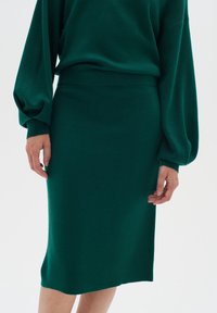 Femme portant une jupe ajustée longueur genou vert foncé et un haut assorti à manches longues bouffantes, debout devant un fond uni.