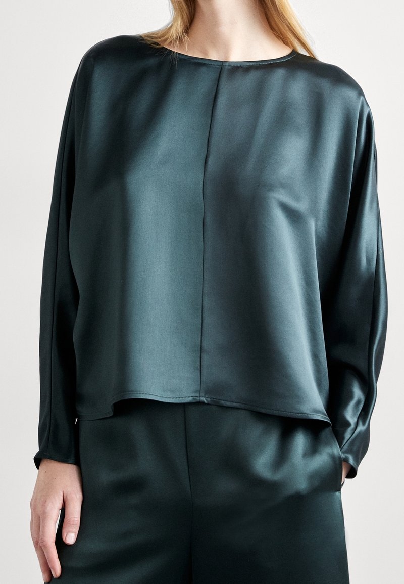 Femme portant un haut en satin vert foncé à manches longues et un pantalon assorti, debout devant un fond uni.