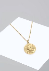 Collier en or avec un pendentif rond présentant le profil détaillé d'un Amérindien portant une coiffe traditionnelle, sur une surface d'exposition blanche.