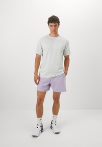 Lichtgrijs shirt met korte mouwen met een gestructureerde afwerking, gecombineerd met lila shorts. Sneakers met zwarte en paarse accenten. Witte achtergrond.