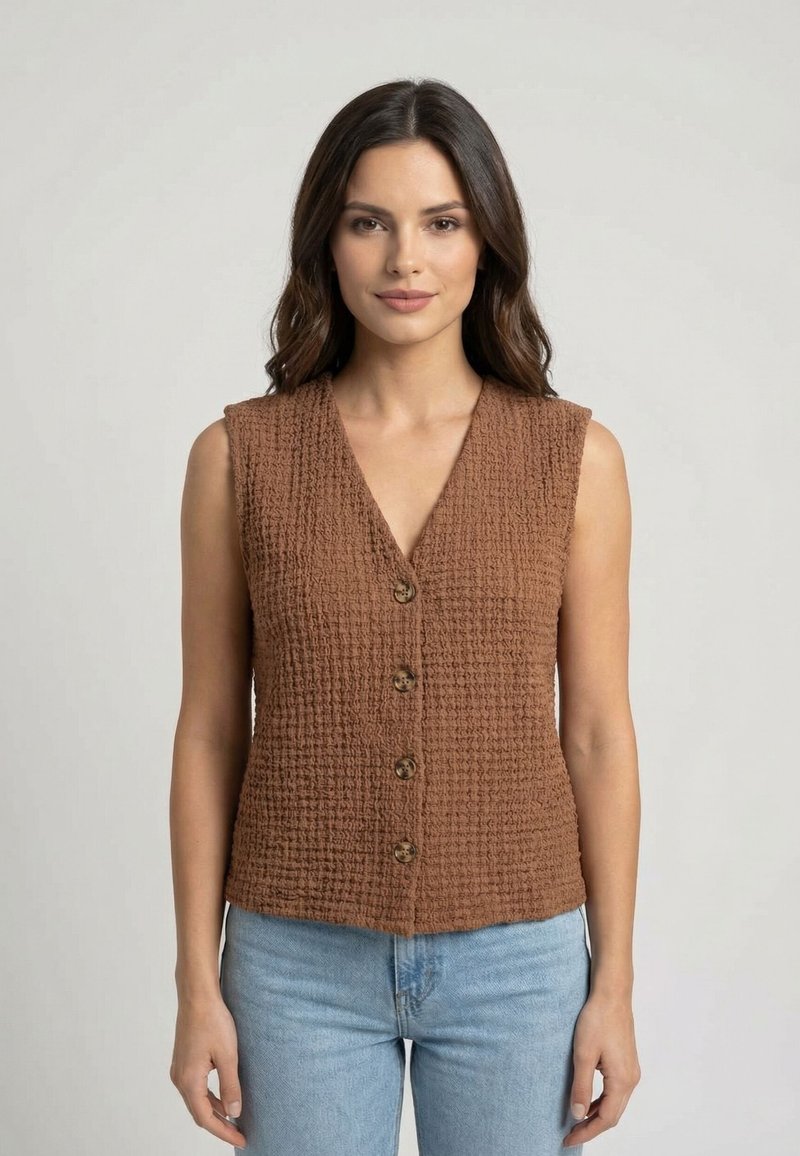 Giovane donna con capelli lunghi e scuri indossa un gilet marrone senza maniche con bottoni e jeans azzurri, in piedi davanti a uno sfondo chiaro uniforme.