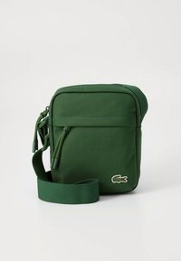 Groene crossbodytas van canvas met ritssluiting, verstelbare riem en een klein geborduurd logo aan de voorkant. Compact en rechthoekig ontwerp.
