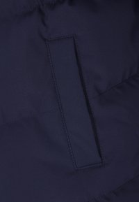 Közeli kép egy tengerészkék steppelt anyagról, amelyen függőleges, varrással körvonalazott hasítékzseb található.