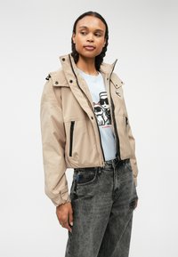 Beige színű, könnyű dzseki cipzárral, állítható kapucnival és fekete cipzár részletekkel. Világoskék grafikás pólóval és szürke farmernadrággal viselve.