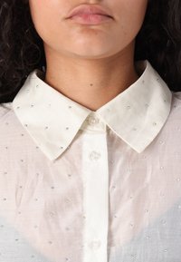 Hvit skjorte med krage og glatt tekstur, med iriserende dekorasjoner jevnt fordelt over stoffet. To knapper ved kragen.