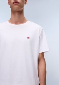 Hvit bomull t-skjorte med rund hals og korte ermer. Har et lite brodert norsk flagg på brystet. Glatt tekstur.
