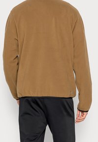 Brun fleecejacka med rund halsringning, elastisk nederkant och muddar. Har en slät textur och minimal logotyp på nederkanten.