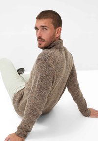 Brauner gestrickter Pullover mit hohem Kragen und strukturiertem Muster. Getragen mit hellen Hosen, sitzt auf einer weißen Oberfläche.
