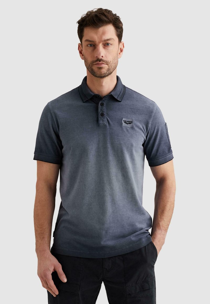 Hombre con una camiseta tipo polo de manga corta azul oscuro con cuello y pantalones cargo negros, de pie con una mano en el bolsillo.