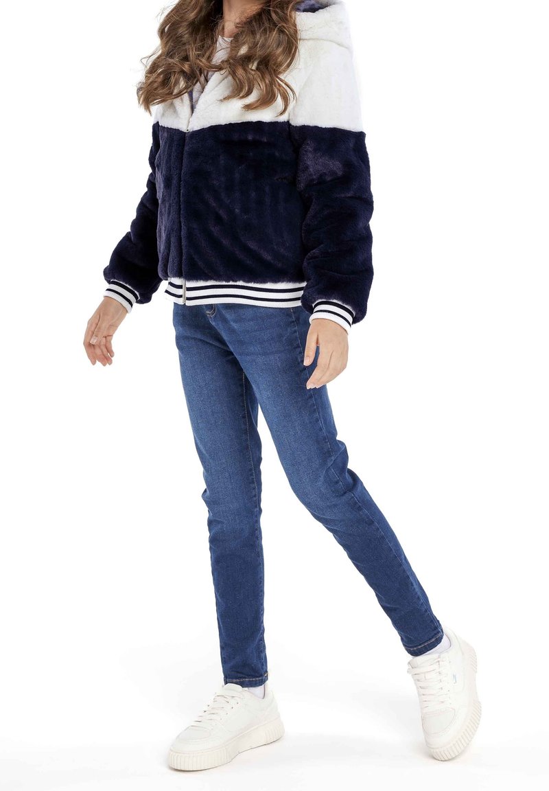 Personne portant une veste pelucheuse bleu marine et blanche, un jean bleu et des baskets blanches, se tenant debout avec une jambe légèrement levée sur un fond blanc.
