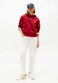 Rode hoodie met wit logo, laag over een roze en wit gestreept shirt. Witte broek en witte sneakers maken de outfit compleet. Casual ontwerp.