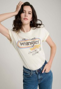 Cremefarbenes Baumwoll-T-Shirt mit retro "Wrangler"-Grafik in Dunkelbraun, Gelb und Blau, kombiniert mit blauen Denim-Jeans.