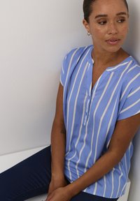 Blouse bleu clair à manches courtes avec des rayures verticales blanches, un col en V avec un détail de bouton, et une texture douce et lisse.