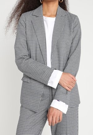 Femme portant un blazer à carreaux noir et blanc et un pantalon assorti sur une chemise blanche à manches longues, bras croisés à la taille.