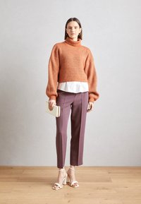 Kraftig orange stickad polotröja lagd över en vit blus, matchad med plommonfärgade byxor och vita blockklackade sandaler, hållande en kuvertväska.