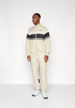 Beige tracksuit with navy and white stripes, zip-up jacket, elastic cuffs, and matching tapered pants. Worn with white sneakers.