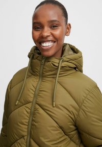Giacca imbottita verde oliva con cappuccio, dotata di zip frontale, coulisse e texture trapuntata. Modello sorridente che mostra la vestibilità.