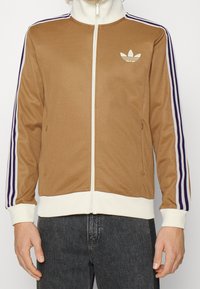 Brun zip-up jacka med vita detaljer, med lila-randiga ärmar, hög krage och en liten vit logotyp på bröstet.