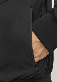 Czarna kurtka z boczną kieszenią na zamek. Materiał jest gładki, z lekko teksturowanym mankietem, co podkreśla elegancki i nowoczesny design.