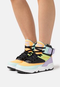 Vysoké tenisky s kombinací oranžové, černé, mátově zelené a levandulové. Mezi rysy patří pásek, texturované panely a kontrastní podešev.