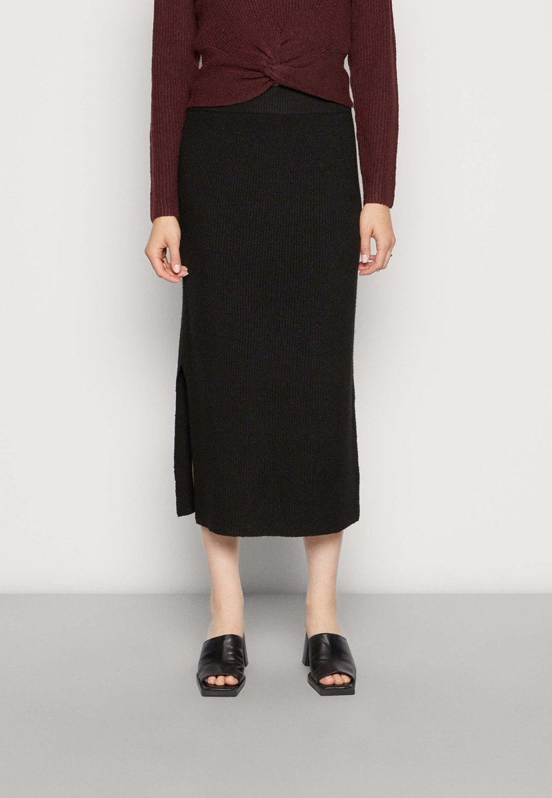 Saia midi preta de malha canelada com uma fenda lateral. Combinada com mules pretos de salto bloco aberto na frente e uma blusa de mangas longas cor bordeaux.